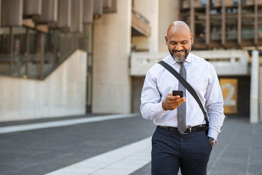 An older professional Black man walking while smiling down at his phone. Representing how our online continuing education for therapists can be done on the go! Explore our courses today. 