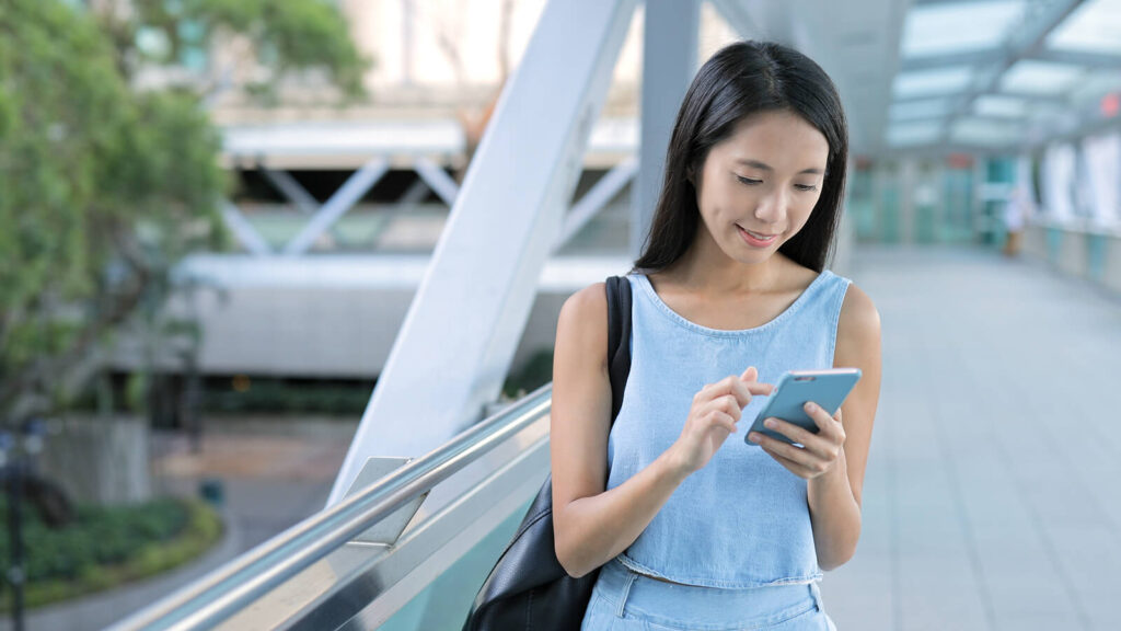 An Asian american woman smiling while scrolling on her phone & walking. Representing how professional development can help therapists reignite their passion. Discover online therapist continuing education by exploring our courses.