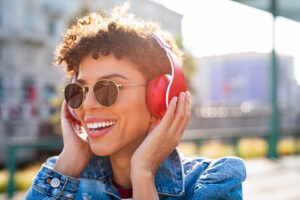 A woman holding red headphones to her ears while smiling outside. 
